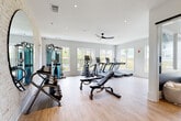 This single-story fitness center is thoughtfully designed to support a comprehensive workout experience, combining modern aesthetics with a well-curated equipment selection. The space is divided into distinct functional zones, each designed to accommodate different training modalities.

The main cardio area features multiple treadmills and an elliptical machine positioned along a bank of large windows that frame outdoor views of the surrounding greenery, flooding the space with natural light. A ceiling fan overhead promotes airflow throughout the room.

Adjacent to the cardio zone, a strength training area is anchored by a multi-station cable machine and a full dumbbell rack set against a distinctive whitewashed brick accent wall. A large round mirror reflects the open layout and enhances the sense of space. A sleek black-framed glass door serves as a striking architectural detail that separates zones while maintaining visual connectivity.

A dedicated functional training area includes adjustable benches, a wall-mounted television, and a rack stocked with medicine balls, resistance bands, and stability balls of varying sizes. A sliding barn door adds a modern industrial touch to this section.

Throughout the space, light wood-toned flooring ties the zones together, contributing to the bright, clean, and motivating ambiance of the facility.