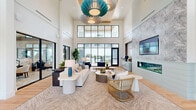 Located in Clermont, FL, this single-floor living space makes a striking first impression with its dramatic double-height ceilings and expansive floor-to-ceiling black-framed windows that flood the interior with natural light. The open-concept layout is anchored by light-toned hardwood flooring that runs throughout, lending warmth and continuity to the space. A statement teal chandelier serves as a bold focal point overhead, complementing the coastal-inspired color palette of whites, blues, and natural textures. The living area is thoughtfully arranged with a plush white sofa, woven area rug, and organic-shaped coffee tables, creating an inviting yet polished atmosphere. Casual seating vignettes featuring navy swivel chairs and round bistro tables offer additional gathering spots, ideal for conversation or leisure. A built-in beverage station with light blue cabinetry and a mosaic tile backsplash adds a refined, functional touch to one corner of the room. Large glass doors open directly to an outdoor terrace, where views of palm trees and a pool area are visible, reinforcing the indoor-outdoor connection. Lush potted greenery and sculptural décor elements further enhance the fresh, resort-inspired ambiance of this airy and welcoming space.