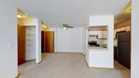 This single-floor residence features an open-concept layout that connects the living area and kitchen, creating a seamless and functional flow throughout the space. The living area is finished with neutral wall-to-wall carpeting and white walls, lending a clean and versatile backdrop suitable for a variety of furnishing styles. A ceiling fan with a light fixture is centrally mounted on the textured white ceiling, providing both ventilation and ambient lighting.

The kitchen is positioned to the side and opens to the living area via a raised breakfast bar with a light-toned countertop, making it ideal for casual dining or entertaining. The kitchen is equipped with a stainless electric range, an overhead range hood, and ample upper cabinetry finished in a warm wood tone. The countertops feature a subtle speckled pattern that complements the overall neutral palette.

A built-in shelving unit is integrated into the wall near the entry, offering practical storage or display space. The front entry door is finished in a natural wood tone, adding warmth to the otherwise light interior. White baseboards frame the perimeter, tying together the clean, consistent aesthetic present throughout the space.