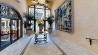 Located at 725 S Bixel St in downtown Los Angeles, this sophisticated lobby space exudes a refined Mediterranean aesthetic that makes an immediate impression upon entry. The grand foyer is anchored by soaring arched windows and doors adorned with intricate wrought-iron scrollwork, allowing natural light to pour in and illuminate the travertine tile flooring below, which features elegant inlaid border detailing. A decorative wrought-iron center table topped with a marble surface and flanked by lush potted palms and floral arrangements creates a striking focal point at the heart of the entry hall. A large tapestry with a richly detailed landscape scene graces one wall, adding an artistic and classical dimension to the space. The warm, golden-toned plaster walls lend a Mediterranean warmth throughout. A curved reception desk with rich wood paneling and ornate detailing serves as a welcoming point for visitors. The lobby also features a dedicated elevator bay with warm marble surrounds, providing seamless vertical access. Staircase elements with curved marble steps and elegant black wrought-iron handrails add to the architectural grandeur. Recessed lighting and wall sconces complement the overall ambiance, creating a polished and welcoming environment.