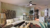 This well-designed single-floor space showcases a modern, open-concept layout that effortlessly blends the living, dining, and kitchen areas into one cohesive environment. The living area is anchored by a sleek, low-profile sofa in a neutral cream tone, complemented by a bold patterned area rug in rich earth tones of brown, red, and black. A rectangular wood-slab coffee table serves as a functional centerpiece, while decorative accents add character throughout. A striking ceiling fan with a built-in light fixture enhances both airflow and ambiance overhead. One wall features large, textured paneling in a soft sage-green tone, creating a refined architectural backdrop, alongside a decorative branch wall art piece that adds a natural, organic element. A tall arc floor lamp provides warm, ambient lighting to the space. The dining area transitions naturally from the living room, featuring a high bistro-style table paired with dark wooden bar stools, ideal for casual dining. A lush indoor plant adds a vibrant pop of greenery. The kitchen, visible in the background, is equipped with stainless steel appliances including a refrigerator and wall oven, dark cabinetry, and light-toned flooring, offering both functionality and style throughout.