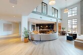 Located at 704 W Tremont Ave in Charlotte's Dilworth neighborhood, this generously sized lobby and hallway space offers a striking first impression through its modern architectural design and open-concept layout. The space is defined by soaring double-height ceilings that flood the interior with natural light through expansive floor-to-ceiling windows and a dramatic glass-paneled upper mezzanine facade. Wide-plank light hardwood flooring runs throughout, lending warmth and continuity across the entire level.

At the heart of the space sits a curved sectional sofa anchored by a patterned area rug, creating an inviting lounge area ideal for gathering. Adjacent to the lounge, a fully equipped kitchen and bar area features dark cabinetry, pendant lighting, and ample seating at the counter. A dining zone with wooden chairs and round tables extends naturally from this hub. A dedicated mailroom area with built-in parcel and mail lockers provides practical resident services seamlessly integrated into the design.

A grand staircase with black metal railings and glass balustrades serves as a bold architectural focal point, connecting to the upper level. Concrete entry steps with iron handrails at the main entrance add an industrial contrast to the refined interior. The overall ambiance balances contemporary sophistication with welcoming comfort, making this lobby a true centerpiece of the building.