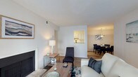 This single-story space presents an open, flowing layout that seamlessly connects the main living area, dining space, and kitchen. The living room serves as the central hub, anchored by a classic fireplace and complemented by warm hardwood-style flooring that extends throughout. Neutral white walls and a textured ceiling create a bright, clean backdrop, while ample lighting enhances the airy atmosphere.

A defining architectural feature is the pass-through window connecting the kitchen to the living area, offering both functional convenience and an open sightline. The kitchen, visible through the pass-through, features white cabinetry and maintains a tidy, efficient layout.

The dining area flows naturally from the living room and is illuminated by a stylish chandelier, adding a touch of elegance to the space. The open-concept arrangement between the living and dining areas promotes a sense of spaciousness and ease of movement.

Overall, the space offers a balanced combination of comfort and functionality, with cohesive finishes, consistent flooring, and a well-connected layout that makes it both practical and inviting for everyday living.