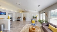 This single-story space offers a thoughtfully designed open floor plan that promotes a seamless flow between living and functional areas. Light wood-look flooring extends throughout, complementing the neutral gray and white color palette that gives the interior a clean, airy feel.

The living area is generously proportioned, featuring vaulted and angled ceilings that enhance the sense of openness. Multiple windows dressed with horizontal blinds allow abundant natural light to fill the room, and views of the outdoors add a refreshing connection to the exterior. The space comfortably accommodates a full seating arrangement centered around a woven ottoman coffee table.

A set of glass-paned French doors opens to a dedicated fitness room, equipped with exercise machines and separated neatly from the main living space while remaining visually accessible.

The kitchen is designed for both function and social engagement, featuring white shaker-style cabinetry, stainless steel appliances, and a wide peninsula countertop lined with industrial-style bar stools—ideal for casual dining or entertaining. Open shelving adds display space, and the kitchen's elevated ceiling contributes to its bright, open character.

Adjacent areas offer additional flexibility, with glimpses of a dining space visible beyond the main living zone, completing a well-rounded and versatile interior.