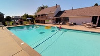 This community amenity space offers residents a comprehensive set of shared facilities designed for both recreation and everyday convenience. At the heart of the outdoor area is a generously sized swimming pool with crystal-clear blue water, framed by a broad concrete sun deck. The deck is well-furnished with multiple lounge chairs and shaded seating areas, providing ample space for relaxing under open skies. A white pergola anchors one end of the patio, adding architectural character, while lush landscaping and iron fencing define the perimeter. The pool area is further complemented by views of the surrounding community buildings and mature trees.

Transitioning indoors, the laundry facility is bright and efficiently laid out, featuring multiple commercial-grade washers and dryers, a folding table, and clean, light-toned walls with overhead fluorescent lighting. The space is accessed through a distinctive dark-paneled entry door with a small window, lending a practical yet tidy feel. Adjacent to the laundry room, a dedicated corridor provides access to a row of individual storage units, each numbered for easy identification and illuminated by recessed ceiling lights. Together, these amenities form a functional and well-maintained shared space that supports the day-to-day needs of the community.