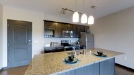 Located at 6634 Riverside Drive in Dublin, Ohio, this single-floor space offers a thoughtfully designed interior with a strong emphasis on the kitchen as a central feature. The kitchen is appointed with rich, dark espresso-toned cabinetry that extends to the upper walls, providing generous storage throughout. Stainless steel appliances, including a refrigerator, electric range, and over-the-range microwave, lend a sleek, contemporary feel to the space.

A expansive granite-topped island serves as both a functional workspace and a casual dining area, set with place settings that highlight its entertaining potential. The island's light-toned granite countertops offer a striking contrast to the darker cabinetry. Overhead, a trio of pendant lights hangs above the island, complementing the track lighting mounted on the ceiling to ensure the kitchen is well-lit throughout. The neutral wall tones and clean lines throughout the space contribute to a modern, cohesive aesthetic that is both functional and visually appealing.