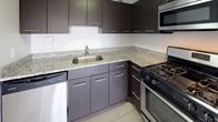 This single-floor unit is defined by its polished parquet wood flooring, which runs consistently throughout the living, bedroom, and connecting areas, lending warmth and a classic character to the space. The open living area is particularly notable for its floor-to-ceiling sliding glass doors, which flood the room with natural light and frame urban views of the surrounding cityscape. A baseboard heating unit runs along the exterior wall, ensuring year-round comfort.

The bedroom is a well-proportioned, quiet room featuring a large window fitted with blinds, a wall-mounted radiator unit, and a flush-mount ceiling fixture. Natural light enters freely, making the room feel airy and comfortable.

The kitchen has been updated with dark espresso-toned cabinetry extending from floor to ceiling, light granite countertops, a stainless steel undermount sink, and stainless steel appliances including a gas range, built-in microwave, and dishwasher. Light-toned tile flooring grounds the space and contrasts cleanly with the cabinetry.

The bathroom maintains a clean, classic aesthetic with white subway tile walls, a soaking tub with shower, chrome fixtures, and a recessed medicine cabinet mirror. The overall ambiance of the unit is bright, functional, and well-maintained throughout.