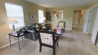 This single-story space centers around a well-appointed living area finished in warm, neutral tones that create an inviting atmosphere. The living room is furnished with a plush, dark gray sofa accented with decorative throw pillows, complemented by a pair of accent chairs — one upholstered in a polka-dot pattern and another with a classic cushioned design. A rectangular coffee table anchors the seating arrangement atop wall-to-wall carpeting that extends throughout the space, adding warmth underfoot. Natural light filters in through multiple windows fitted with white blinds, brightening the room without overwhelming it. A tall decorative plant adds a touch of greenery and visual height to the corner. The living area opens toward an adjacent room through a wide doorway, suggesting a generous, flowing floor plan. A pass-through window or open cutout connects to what appears to be a kitchen area, enhancing the open-concept feel of the layout. Framed wall art and a table lamp contribute to the overall polished, homey ambiance of the space.