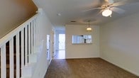This two-story space offers a functional and straightforward layout designed for comfortable everyday living. The main level showcases a carpeted living area beneath a ceiling fan that provides both light and airflow. A decorative pendant light with a brass fixture hangs nearby, adding a subtle accent to the space.

A prominent pass-through window connects the living area to the kitchen, offering a glimpse of white cabinetry and appliances, including a refrigerator. This architectural detail creates an open, communicative feel between the two areas without fully removing the wall separation.

A staircase with white-painted balusters is positioned along one side of the main level, providing access to the upper floor. Near the base of the stairs, a small built-in shelf adds a practical storage element.

Toward the rear of the main level, a doorway leads to a tiled entry or transitional space that opens to a glass-paneled exterior door, allowing natural light to pour into the interior. The walls throughout are painted in a neutral white tone, complementing the light-colored trim and contributing to a clean, bright overall ambiance. The textured ceiling is consistent throughout the visible areas.