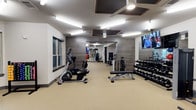 This fitness center offers a thoughtfully designed workout space that balances functionality with a modern aesthetic. The room features neutral-toned flooring that provides a clean, durable surface throughout. One wall is clad in wood-textured paneling, adding a warm, contemporary contrast to the otherwise crisp white walls and ceiling. A large mirror at the far end of the room visually expands the space and allows users to monitor their form during workouts.

Lighting is provided by a combination of recessed ceiling fixtures and ceiling-mounted fans with integrated lighting, ensuring the space remains bright and well-ventilated. A wall-mounted flat-screen television is positioned for easy viewing during exercise sessions.

The equipment layout is organized and spacious, featuring cardio machines including a recumbent bike and elliptical, a dedicated free weights area with a tiered dumbbell rack, and a flat weight bench for strength training. Additional equipment is visible toward the rear of the room. Windows along one wall allow natural light to supplement the interior lighting, contributing to an open, energizing atmosphere well-suited for daily fitness routines.