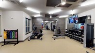 This fitness center offers a thoughtfully designed workout space that balances functionality with a modern aesthetic. The room features neutral-toned flooring that provides a clean, durable surface throughout. One wall is clad in wood-textured paneling, adding a warm, contemporary contrast to the otherwise crisp white walls and ceiling. A large mirror at the far end of the room visually expands the space and allows users to monitor their form during workouts.

Lighting is provided by a combination of recessed ceiling fixtures and ceiling-mounted fans with integrated lighting, ensuring the space remains bright and well-ventilated. A wall-mounted flat-screen television is positioned for easy viewing during exercise sessions.

The equipment layout is organized and spacious, featuring cardio machines including a recumbent bike and elliptical, a dedicated free weights area with a tiered dumbbell rack, and a flat weight bench for strength training. Additional equipment is visible toward the rear of the room. Windows along one wall allow natural light to supplement the interior lighting, contributing to an open, energizing atmosphere well-suited for daily fitness routines.