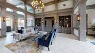 Located at 385 N Godley Station Blvd in Pooler, Georgia, this sophisticated single-floor clubhouse and leasing lobby makes a strong first impression with its grand entrance. A rich mahogany door with decorative leaded glass sidelights and transom windows welcomes visitors into a well-appointed foyer, where large-format tile flooring, a bold blue area rug, and flanking wall sconces set a polished tone.

Structural columns with detailed millwork and wainscoting define the transition from the entry into the expansive main lounge. This central gathering space commands attention with soaring double-height ceilings, a statement chandelier, and an abundance of natural light. Built-in cabinetry with display shelving lines the walls, providing both function and visual depth, while plush navy velvet seating and layered area rugs contribute warmth and elegance.

A dedicated seating alcove offers an intimate setting beneath tall windows that frame sweeping views of the outdoor pool courtyard and surrounding community. Contemporary abstract artwork and velvet accent chairs in deep navy complete the curated aesthetic. Throughout the space, a cohesive palette of warm greige tones, taupe millwork, and gold accents creates a refined and inviting ambiance well-suited for both welcoming residents and hosting community gatherings.