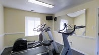 This single-floor fitness center provides a practical and well-equipped space for cardiovascular exercise and general workouts. The room features a selection of cardio machines, including a stationary bike, an elliptical trainer, and a treadmill, offering users a variety of exercise options. A wall-mounted flat-screen television is positioned for easy viewing during workouts, enhancing the overall experience. Large mirrored panels line one wall, visually expanding the space and allowing users to monitor their form. Natural light filters into the room through multiple windows fitted with horizontal blinds, creating a bright and energizing atmosphere. The walls are painted in a soft, neutral yellow-green tone, contributing to a clean and inviting environment. Overhead fluorescent lighting ensures the space remains well-lit at all times. The floor is finished with a durable, dark-toned carpet suitable for fitness use. Ventilation is provided through ceiling vents, maintaining a comfortable environment during exercise. Overall, this fitness center is designed to be functional and accessible, providing essential workout amenities in a well-maintained, light-filled setting.