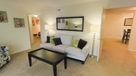This single-floor residence centers around a welcoming living room designed with a clean, contemporary aesthetic. The space is finished with wall-to-wall carpeting in a warm neutral tone, contributing to a comfortable and inviting atmosphere. Smooth, light-colored walls provide a bright backdrop throughout.

The living room is furnished with a generously sized white sectional sofa accented by contrasting black and green decorative pillows, paired with a sleek black rectangular coffee table. A large framed mirror mounted on the main wall adds depth and reflects ambient light, enhancing the sense of openness. Two matching floor lamps with metallic bases flank the seating area, providing layered lighting.

A hallway visible from the living room leads to additional areas of the home, including what appears to be a bedroom featuring its own decorative touches. A separate doorway on the opposite side provides access to another room, suggesting a practical and connected layout. The overall design balances functionality with a modern, coordinated style, making the space feel polished and move-in ready.