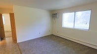 This single-floor space presents a straightforward, functional layout centered around a carpeted room finished in neutral tones throughout. The room features light beige wall-to-wall carpeting that extends across the full floor area, contributing to a quiet and understated atmosphere. The walls are painted in a clean, off-white tone, keeping the interior bright and versatile.

Climate control is well-covered, with both a window-mounted air conditioning unit and a baseboard heating system along the exterior wall, ensuring year-round comfort regardless of season. A single window fitted with horizontal blinds allows natural light to filter into the space, providing a connection to the outdoors while maintaining privacy options.

The room's simplicity and clean lines make it highly adaptable for a variety of uses, whether as a living area or bedroom. Electrical outlets are positioned along the baseboards for convenient placement of furnishings and appliances. The overall ambiance is modest and practical, offering a blank canvas that can be personalized to suit individual needs and preferences.