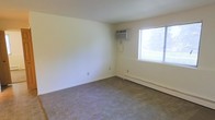 This single-floor space presents a straightforward, functional layout centered around a carpeted room finished in neutral tones throughout. The room features light beige wall-to-wall carpeting that extends across the full floor area, contributing to a quiet and understated atmosphere. The walls are painted in a clean, off-white tone, keeping the interior bright and versatile.

Climate control is well-covered, with both a window-mounted air conditioning unit and a baseboard heating system along the exterior wall, ensuring year-round comfort regardless of season. A single window fitted with horizontal blinds allows natural light to filter into the space, providing a connection to the outdoors while maintaining privacy options.

The room's simplicity and clean lines make it highly adaptable for a variety of uses, whether as a living area or bedroom. Electrical outlets are positioned along the baseboards for convenient placement of furnishings and appliances. The overall ambiance is modest and practical, offering a blank canvas that can be personalized to suit individual needs and preferences.