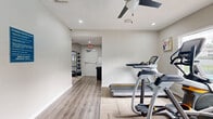 Located in Peachtree Corners, GA, this fitness center is organized across two interconnected exercise rooms, offering a comprehensive workout environment in a compact, efficiently utilized space. The layout separates cardio and strength training areas, allowing users to move seamlessly between zones.

The first room features a standout heartbeat mural painted in blue along the wall, adding an energetic and motivating atmosphere. It is equipped with indoor cycling bikes, a weight bench, and a rack of medicine and stability balls in various sizes. Patterned carpet tiles cover the floor, and a ceiling fan provides ventilation throughout the space.

The second room transitions to light wood-look flooring and hosts cardio machines including a treadmill and an elliptical trainer, positioned near a large window that allows natural light and views of an outdoor pool area. A multi-function cable machine and a dumbbell rack with a wall-mounted flat-screen TV complete the strength training setup. This room also benefits from a ceiling fan and recessed lighting.

Both rooms are finished with neutral gray-toned walls, clean white trim, and recessed lighting, creating a bright and focused workout environment. The glass entry door and clearly marked exit ensure easy accessibility throughout the facility.
