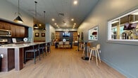 Located at 6741 W 135th St in Overland Park, Kansas, this single-floor amenity center offers a generous and thoughtfully designed space for recreation, fitness, and socializing. The layout encompasses a modern fitness center, a versatile social lounge, a game room, and a fully equipped kitchen area.

The fitness center is a standout feature, boasting high ceilings with an exposed industrial-style roof structure, ceiling fans, and expansive floor-to-ceiling windows that flood the space with natural light and outdoor views. It is outfitted with a comprehensive array of equipment including treadmills, ellipticals, strength machines, free weights, exercise balls, and a dedicated stretching and functional training area with rubberized flooring and wall mirrors.

The social lounge presents a warm, inviting atmosphere with soft blue-gray walls, luxury vinyl plank flooring, and eclectic contemporary décor including curated artwork. Flexible seating arrangements include lounge sofas, dining tables, and high-top bar seating, all centered around a wall-mounted television and decorative stacked wood feature wall. A full kitchen with dark wood cabinetry, granite countertops, and bar stools supports entertaining and gatherings.

A dedicated game room features a pool table, shuffleboard, and billiard-themed wall art. A carpeted corridor with dual handrails and art displays connects the various areas of this well-rounded community facility.