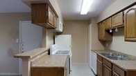 This single-story space centers around a practical kitchen that offers a straightforward and functional layout. The kitchen is equipped with white appliances, including a freestanding electric range, a refrigerator, a built-in microwave, and a dishwasher, providing all essential cooking and meal preparation amenities. Dark wood cabinetry lines both sides of the kitchen, offering ample upper and lower storage space. The countertops feature a light-toned laminate surface that contrasts with the warm brown tones of the cabinetry. A double sink is positioned along one wall, complemented by additional cabinet storage beneath. The flooring throughout appears to be a light-colored tile, contributing to a clean and easy-to-maintain environment. The textured ceiling is illuminated by a fluorescent overhead light fixture, providing adequate task lighting across the workspace. The neutral beige wall tones keep the space feeling cohesive and understated. A corridor visible at the rear of the kitchen suggests a connection to additional rooms within the unit, indicating a straightforward and efficiently organized floor plan overall.