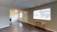 This single-floor unit offers a clean, functional layout designed for comfortable everyday living. The main living area is finished with rich, dark hardwood-style flooring that runs throughout, providing a warm contrast to the neutral beige walls and crisp white trim. A textured ceiling adds a classic touch overhead, while a simple flush-mount light fixture provides ambient illumination. Natural light flows generously into the space through multiple windows fitted with white horizontal blinds, offering views of the surrounding exterior. A wall-mounted PTAC unit situated beneath the large front-facing window ensures efficient heating and cooling throughout the year. The entryway is defined by a solid white door accompanied by a secondary window, creating a bright and welcoming point of entry. The open design of the living space allows for flexible furniture arrangement, making it easy to personalize. The overall ambiance is bright, clean, and move-in ready, with a straightforward aesthetic that provides a neutral canvas for any interior style. This well-maintained space balances simplicity and practicality, making it a comfortable and inviting place to call home.