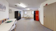 Located at 2750 S 38th St in Grand Forks, North Dakota, this community room serves as a practical and welcoming shared space for residents and visitors. The room is finished with neutral-toned walls and wall-to-wall carpeting that lends a warm, comfortable feel underfoot. Overhead fluorescent lighting ensures the space is well-lit throughout.

The layout accommodates multiple seating arrangements, including a modern dining-style table with sleek white chairs featuring wood-toned legs, ideal for casual gatherings or meetings. A secondary folding table with additional white chairs offers a dedicated spot for reading or light activities, complemented by a small collection of books displayed on the table surface. Framed artwork and small decorative pieces adorn the walls, adding a touch of character to the otherwise functional environment.

A snack vending machine and a Coca-Cola beverage vending machine are conveniently positioned within the room, providing easy access to refreshments. Windows along the walls allow for natural light to filter in, brightening the interior. A corridor connects the community room to the broader building, with wood-paneled doors offering easy access. The overall ambiance is clean, practical, and inviting — well-suited for community interaction and everyday use.