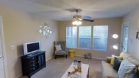 This single-floor space centers around a welcoming living room designed for both comfort and style. The room features plush carpeted flooring and neutral-toned walls that create a warm, inviting atmosphere. A multi-blade ceiling fan with an integrated light fixture serves as a functional focal point overhead, complemented by a textured popcorn ceiling. Natural light fills the space through a trio of windows dressed with horizontal blinds, offering views of surrounding greenery outside.

The living area is thoughtfully arranged with a mix of seating options, including a classic wingback chair in a soft gray upholstery paired with a bold yellow accent stool, adding a pop of color to the neutral palette. A decorative cluster of circular mirrors adorns one wall, enhancing the sense of depth and light in the room. A dark wood media console with drawer storage provides practical entertainment space, while a white coffee table anchors the seating area. Decorative lanterns and driftwood accents add a relaxed, coastal-inspired touch, contributing to the overall cozy and casual ambiance of the space.