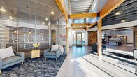 Located at 1331 Crossings Blvd in Shakopee, Minnesota, this single-floor community space offers a generously sized, open-concept layout designed with a contemporary industrial aesthetic. The space makes an immediate impression with its soaring ceilings, exposed steel trusses, and visible ductwork, balanced by the warmth of natural wood beams and richly textured stacked-stone accent walls. Large-format tile flooring in the main entry area transitions seamlessly into hardwood and patterned carpet throughout the lounge zones, creating distinct yet connected areas within the open floor plan.

The welcoming entrance features glass-paneled partitions, a bold barn-style sliding door, and generous glazing that floods the interior with natural light. A spacious living area is anchored by a floor-to-ceiling stacked-stone fireplace, complemented by oversized windows offering views of the surrounding neighborhood. Comfortable sectional seating with vibrant accent pillows adds a lively, residential feel to the communal lounge.

A bar-height counter seating area with pendant lighting and direct access to an outdoor patio reinforces the space's social character. Colorful artwork and carefully curated furnishings contribute to an energetic yet inviting ambiance, making this a well-appointed gathering space suited for both relaxed lounging and community engagement.
