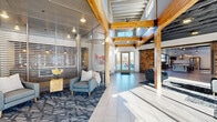 Located at 1331 Crossings Blvd in Shakopee, Minnesota, this single-floor community space offers a generously sized, open-concept layout designed with a contemporary industrial aesthetic. The space makes an immediate impression with its soaring ceilings, exposed steel trusses, and visible ductwork, balanced by the warmth of natural wood beams and richly textured stacked-stone accent walls. Large-format tile flooring in the main entry area transitions seamlessly into hardwood and patterned carpet throughout the lounge zones, creating distinct yet connected areas within the open floor plan.

The welcoming entrance features glass-paneled partitions, a bold barn-style sliding door, and generous glazing that floods the interior with natural light. A spacious living area is anchored by a floor-to-ceiling stacked-stone fireplace, complemented by oversized windows offering views of the surrounding neighborhood. Comfortable sectional seating with vibrant accent pillows adds a lively, residential feel to the communal lounge.

A bar-height counter seating area with pendant lighting and direct access to an outdoor patio reinforces the space's social character. Colorful artwork and carefully curated furnishings contribute to an energetic yet inviting ambiance, making this a well-appointed gathering space suited for both relaxed lounging and community engagement.