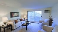 This single-floor residence offers a thoughtfully designed layout that balances comfort and functionality. The living room serves as the heart of the space, featuring plush wall-to-wall carpeting that adds warmth underfoot. A generously sized sliding glass door floods the room with natural light while framing a serene view of mature, lush trees beyond a private balcony — creating a seamless connection between the interior and the outdoors. The balcony itself provides an inviting spot to enjoy the greenery. The living room's neutral color palette, highlighted by crisp white walls and a textured ceiling, creates a clean and airy ambiance that is easy to personalize. A wall-mounted air conditioning unit ensures year-round comfort. The space is versatile and well-proportioned, offering ample room for a full seating arrangement, making it ideal for both relaxing and entertaining. Overall, this residence combines practical amenities with a tranquil, nature-inspired atmosphere, making it a comfortable and inviting place to call home.
