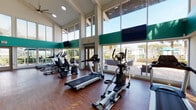 This fitness center is designed to provide a bright, open environment for exercise and wellness. The main cardio area is defined by dramatic double-height ceilings and expansive floor-to-ceiling windows that wrap around the space, flooding the interior with natural light and offering unobstructed views of the surrounding outdoor patio and landscaping. Large ceiling fans circulate air throughout the room, complementing the airy feel created by the soaring architecture. The hardwood-style flooring anchors the space and provides a clean, modern aesthetic. Multiple wall-mounted flat-screen televisions are positioned for visibility from the cardio equipment, which includes treadmills, elliptical trainers, and stationary bikes arranged to take full advantage of the outdoor views. Bold teal and green accent bands run along the upper interior walls, adding a distinctive visual element to the design. French-style glass doors provide direct access to an outdoor patio area. A secondary, lower-ceilinged studio room connects to the main floor and features patterned carpet, recessed lighting, a wall-mounted television, and a freestanding punching bag, making it suitable for stretching, strength training, or group fitness activities. Glass partition walls and sliding doors maintain an open connection between the two areas.