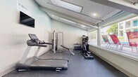 This fitness center is a thoughtfully designed workout space that maximizes functionality within a compact footprint. The room features a sloped, beamed ceiling with recessed lighting and skylights that flood the interior with natural light, creating a bright and energizing atmosphere. Large floor-to-ceiling windows line one side of the space, offering views of the adjacent outdoor pool and common areas, further enhancing the open, airy feel of the room.

The fitness center is equipped with a variety of cardio and strength training equipment, including Spirit-brand treadmills, an elliptical machine, a stationary bike, a multi-function cable weight machine, and an adjustable workout bench. A wall-mounted flat-screen television is positioned for easy viewing during workouts. The durable dark rubber flooring provides a safe, non-slip surface suited for a range of exercise activities.

The interior is finished with clean, neutral-toned walls in a soft sage and white palette, contributing to a calm and focused workout environment. A glass-paneled exterior wall and dedicated entry door provide direct access to the outdoor amenity areas. The space is also equipped with an HVAC vent and emergency exit signage, reflecting attention to comfort and safety standards throughout.