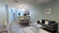 This single-floor residence offers a thoughtfully designed open-concept layout that connects the living, dining, and kitchen areas into one cohesive and welcoming space. The interior is finished with wide-plank hardwood-style flooring that runs throughout, anchoring the modern aesthetic and adding warmth underfoot. The living area is comfortable and well-proportioned, featuring a contemporary sofa set paired with a sleek, mirrored glass coffee table and a plush area rug that adds texture to the space. The dining area sits centrally within the open floor plan, accommodating a four-seat table beneath a striking cluster of cylindrical pendant lights that serve as a focal point and provide warm, direct illumination. Adjacent to the dining space, a kitchen with dark cabinetry and a raised breakfast bar creates a natural division between the cooking and living zones while maintaining the open feel. The soft gray-toned walls complement the neutral furnishings and allow natural and ambient light to reflect evenly across the space, contributing to a bright and airy atmosphere. Recessed ceiling vents and a well-planned layout speak to the functional design of this residence. A hallway leading off the main area provides access to additional rooms, completing this clean, modern living space.