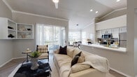 This single-floor residence offers a thoughtfully designed open-concept layout that seamlessly connects the living, dining, and kitchen areas. The living room is a standout space, highlighted by soaring vaulted ceilings with crown molding, herringbone-patterned hardwood-style flooring, and a sliding glass door that floods the room with natural light and provides access to an outdoor balcony. A pass-through kitchen features white cabinetry with black hardware, a gold-toned faucet, stainless steel appliances including a range and microwave, and light-toned countertops. Adjacent to the kitchen, the dining area is framed by large windows dressed with horizontal blinds, creating a bright and airy atmosphere beneath a modern ring pendant light. The hallway, finished with the same herringbone flooring, connects the main living spaces to the private quarters. The bedrooms are carpeted and serene, with crown molding, ceiling fans, and ample closet space. Two full bathrooms are clean and functional, each featuring a tub-shower combination with white tile surround and herringbone-style flooring. Neutral warm-gray tones carried throughout the space create a cohesive and inviting ambiance.