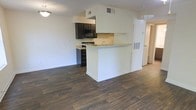 Located at 5639 Old Chapel Hill Rd in Durham, North Carolina, this single-floor space offers a clean, modern layout designed for comfortable everyday living. The open-concept floor plan seamlessly connects the kitchen and living area, creating a bright and airy atmosphere throughout. Rich, dark hardwood-style flooring runs across the main living space, providing a warm contrast to the crisp white walls and trim. The kitchen is outfitted with dark cabinetry featuring sleek hardware, black appliances including a built-in microwave and range, and light-toned countertops. A peninsula-style breakfast bar extends from the kitchen, offering additional counter space and serving as a natural divider between the kitchen and the open living area. Overhead ventilation and recessed fixtures contribute to the functional design. The neutral color palette of white walls and ceilings keeps the space feeling open and versatile. A hallway visible from the main living area leads to additional rooms, suggesting a well-organized and practical floor plan. Overall, the space presents a tidy, move-in-ready environment with contemporary finishes throughout.