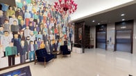 This hotel lobby makes an immediate visual statement through a combination of contemporary design elements and bold artistic choices. The most commanding feature is a large-scale, floor-to-ceiling mural depicting a colorful, diverse crowd of illustrated figures, serving as a dynamic focal point that sets an energetic and welcoming tone. Suspended above the seating area are two dramatic red glass chandeliers, adding a rich pop of color against the bright white ceiling and recessed lighting. The flooring throughout is polished light-toned tile, lending an open and clean aesthetic to the space. A seating arrangement of deep navy blue velvet chairs with gold-toned legs is positioned beneath the mural, offering guests a comfortable place to gather. The front desk area is defined by dark paneling with horizontal gold accent striping, paired with a sleek lamp and a dark marble base, providing an elegant and professional check-in experience. Decorative dark shelving with gold trim lines additional walls, complementing the overall color palette. The elevator bank features stainless steel doors framed in rich dark wood paneling, alongside a built-in mailbox wall unit. Gold-accented open shelving and a bellhop cart complete the functional yet stylish back-of-lobby area, reinforcing the space's blend of practicality and refined hospitality design.