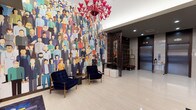 This hotel lobby makes an immediate visual statement through a combination of contemporary design elements and bold artistic choices. The most commanding feature is a large-scale, floor-to-ceiling mural depicting a colorful, diverse crowd of illustrated figures, serving as a dynamic focal point that sets an energetic and welcoming tone. Suspended above the seating area are two dramatic red glass chandeliers, adding a rich pop of color against the bright white ceiling and recessed lighting. The flooring throughout is polished light-toned tile, lending an open and clean aesthetic to the space. A seating arrangement of deep navy blue velvet chairs with gold-toned legs is positioned beneath the mural, offering guests a comfortable place to gather. The front desk area is defined by dark paneling with horizontal gold accent striping, paired with a sleek lamp and a dark marble base, providing an elegant and professional check-in experience. Decorative dark shelving with gold trim lines additional walls, complementing the overall color palette. The elevator bank features stainless steel doors framed in rich dark wood paneling, alongside a built-in mailbox wall unit. Gold-accented open shelving and a bellhop cart complete the functional yet stylish back-of-lobby area, reinforcing the space's blend of practicality and refined hospitality design.