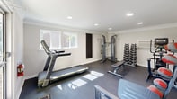 Located at 317 Atwood Dr in Burlington, NC, this dedicated exercise room offers a comprehensive fitness facility within a compact, efficiently organized space. The room features neutral light-gray walls, crown molding, and a dark rubber flooring that is both durable and practical for workout use. Recessed lighting throughout provides bright, even illumination, complemented by natural light streaming in through multiple windows and a glass-panel door that opens to an outdoor view of mature trees and greenery.

The space is thoughtfully equipped with a wide range of fitness equipment, including a Matrix treadmill, an elliptical trainer, a leg press and cable machine, a multi-function cable crossover system, a flat adjustable bench, and a True fitness bench. A full rack of hex dumbbells in graduated sizes provides ample free-weight options, while a set of medicine balls on a dedicated rack adds versatility for functional training. A wall-mounted PTAC unit ensures climate control year-round. Safety guidelines are posted on the wall, and sanitation supplies are conveniently accessible near the entrance. The glass door allows easy outdoor access, enhancing the open and airy feel of this well-appointed fitness space.