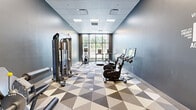 This fitness center offers a thoughtfully designed workout space that maximizes functionality within a compact footprint. The room features cool-toned, slate-blue and charcoal walls that lend a modern, energizing atmosphere, complemented by a distinctive geometric-patterned floor in shades of gray, beige, and white that adds visual dynamism to the space.

A large, multi-pane floor-to-ceiling window anchors one wall, flooding the gym with natural light and providing an open view of the surrounding outdoor greenery — an inviting backdrop for cardio workouts. Recessed ceiling panels with integrated lighting ensure consistent illumination throughout.

The facility is equipped with a diverse range of exercise equipment. Cardio options include multiple treadmills and an elliptical trainer, all positioned to face the expansive window. A pair of stationary cycling bikes with digital screens occupy a dedicated area alongside a motivational "FITNESS" wall graphic. A multi-function cable resistance machine supports a wide variety of strength training exercises. A well-stocked accessory station rounds out the space, featuring stability balls, resistance bands, medicine balls, and foam rollers — ideal for stretching and functional training routines. The overall layout is open and organized, allowing for comfortable movement between stations.