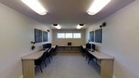 This single-floor computer lab is thoughtfully designed to support focused work and collaborative learning in a clean, organized environment. The room features two long, light wood-toned workstation counters arranged symmetrically along the side walls, each outfitted with desktop computers and ergonomic task chairs, offering ample workspace for multiple users simultaneously. A central aisle provides easy navigation between the two rows, while a additional counter along the back wall offers supplementary workspace or equipment placement.

The space is well-lit by a combination of flush-mount and decorative ceiling light fixtures that distribute even, bright illumination throughout. A horizontal window centered on the back wall allows natural light to filter in, adding a sense of openness to the interior. The neutral-toned walls and light carpeted flooring create a calm, distraction-free atmosphere conducive to productivity.

Decorative elements include a series of geometric, textured wall panels mounted symmetrically on either side of the room, complemented by sculptural starburst wall accents that add visual interest without overwhelming the functional intent of the space. The overall design strikes a balance between practicality and modern aesthetic, making it a versatile and welcoming environment for tech-focused tasks.