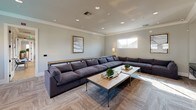 This contemporary single-floor lounge and media room is thoughtfully designed for both relaxation and entertainment. The space is anchored by a generously sized L-shaped sectional sofa upholstered in deep charcoal fabric, arranged around a rustic wood-and-metal coffee table accented with decorative greenery. Neutral-toned walls, crown molding, and recessed lighting overhead create a refined yet comfortable atmosphere, while patterned carpet flooring adds warmth and texture underfoot.

The entertainment wall is a standout feature, showcasing multiple large flat-screen televisions mounted above a sleek, dark built-in media console with ample drawer storage. This setup makes the space ideal for watching multiple broadcasts or presentations simultaneously. A bar-height communal table with metal-framed stools is positioned nearby, offering a more casual seating option perfect for gathering or working.

Natural light filters in through a series of horizontal windows positioned along the upper portion of the walls, maintaining privacy while brightening the interior. A floor lamp and framed wall art contribute to the curated, welcoming ambiance. The open layout allows for easy movement throughout the room, making it well-suited as a shared amenity or common area lounge. Overall, the space balances modern design sensibilities with functional, multi-use versatility.