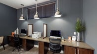 Located in the Baldwin Park neighborhood of Orlando, Florida, this single-floor business center at 4688 New Broad St offers a well-appointed and functional workspace designed with a cohesive, contemporary aesthetic. The space is characterized by deep slate-blue and charcoal wall tones, crisp white trim, and a mix of tile and patterned carpet flooring that defines different zones throughout.

The main workstation area features a long, continuous built-in desk with warm wood cabinetry, accommodating multiple iMac computers and a multifunction printer. Industrial-style polished nickel pendant lights hang overhead, providing focused task lighting. Framed whiteboard panels are integrated into the workspace for added utility.

A semi-private lounge and meeting area offers a more relaxed setting, furnished with a cushioned sofa, a round bistro-style table with chairs, and an additional workstation. Natural light filters in through large windows fitted with wood-toned blinds, lending warmth to the darker color palette.

The corridor connecting the spaces is clean and bright, with large-format tile flooring and glass-paneled doors leading toward a well-lit reception or common area. Curated abstract artwork adorns the walls, adding a sophisticated, gallery-like quality to the environment. Overall, the space blends professional functionality with thoughtful interior design.
