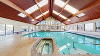 This expansive community amenity facility in Aurora, Colorado offers an impressive array of recreational and social spaces across a generous single-floor footprint. The crown jewel is the stunning indoor pool area, where soaring vaulted ceilings with warm wood beam detailing and large skylights create a bright, airy atmosphere. Floor-to-ceiling windows line the walls, flooding the space with natural light and offering views of the surrounding trees. A hot tub/spa sits adjacent to the pool, and bench seating is provided throughout the area. A hallway entry with polished concrete floors and vaulted wood ceilings serves as a welcoming transition into the facility.

The well-appointed fitness center is equipped with a comprehensive selection of cardio machines—including Cybex ellipticals, stair climbers, and treadmills—alongside a multi-station weight machine, free weights, and benches. Full-wall mirrors and windows overlooking the outdoors enhance the workout environment. A dedicated racquetball court features a classic hardwood floor and clean white walls, providing ample space for play.

The party room includes a comfortable living area with a tiled fireplace, large windows, and natural light, complemented by a functional kitchen featuring wood cabinetry, marble-style countertops, and black appliances—ideal for community gatherings and events.