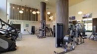 This fitness center offers a well-appointed workout environment designed with both functionality and aesthetics in mind. The space is defined by soaring double-height ceilings that lend an open, airy feel, complemented by large decorative columns clad in rich wood-toned paneling that add an architectural elegance rarely found in a gym setting. Stylish globe pendant lights hang throughout, providing warm, ambient illumination that enhances the overall atmosphere. The walls feature floor-to-ceiling vertical striped accent panels, adding a refined, contemporary touch to the interior design. The facility is outfitted with a wide array of professional-grade strength training machines, strategically arranged to allow ample space between equipment for comfortable movement. Mounted flat-screen televisions are positioned throughout the space, allowing members to stay entertained during their workouts. Light-toned flooring and neutral wall colors contribute to the clean, modern aesthetic. The combination of thoughtful design details, generous vertical space, and a comprehensive equipment selection makes this fitness center a premium amenity that balances a welcoming ambiance with serious workout capability.