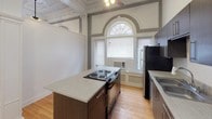 This single-floor space masterfully combines historic architectural character with contemporary finishes. The main living area immediately impresses with its soaring ceilings adorned with ornate plasterwork, decorative columns, and elegant arched doorways that speak to the building's storied past. A coffered ceiling with detailed molding crowns the open living space, while warm hardwood floors run throughout, adding both warmth and continuity.

The kitchen is a functional highlight, featuring a central island with an electric cooktop, dark espresso cabinetry, stainless steel appliances including a full-size refrigerator, and a built-in sink. Tall arched windows with fanlight transoms flood the kitchen with natural light, framing the space with striking architectural detail.

The bedroom is a serene retreat, benefiting from the same generous ceiling height, a ceiling fan with light fixture, and the continuation of gleaming hardwood flooring. An arched architectural feature adds visual interest to the room.

The bathroom is clean and efficiently designed, offering a single-sink vanity with a large mirror, a toilet, and a full bathtub with a shower curtain enclosure, all finished in a crisp, neutral palette.

Throughout the space, the interplay of preserved historic ornamentation and updated modern fixtures creates a distinctive and inviting atmosphere.
