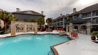 This resort-style pool area offers a welcoming and well-appointed outdoor retreat. The centerpiece is a generously sized swimming pool with striking turquoise water, accented by built-in water features that cascade from stone-clad structures, adding both visual appeal and a relaxing ambiance. The pool is flanked by a broad, tiled sundeck lined with vibrant red lounge chairs and chaise lounges, providing ample space for sunbathing and outdoor relaxation. Towering palm trees are positioned throughout the area, enhancing the tropical atmosphere and providing natural shade. The pool surround features a combination of brick edging and stone masonry columns, contributing to a refined yet casual aesthetic. Multi-story residential buildings with dark siding, pitched roofs, and private balconies frame the backdrop, creating an enclosed and private feel. A gated entry point with a keypad-secured door ensures controlled access to the pool area. Convenient amenities include beverage vending machines positioned near the entrance for easy poolside refreshment. The overall design balances a resort-inspired aesthetic with practical, community-focused features, making this pool area a standout communal space.