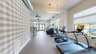 Located on Buckingham Street in Matawan, NJ, this fitness center offers a generously sized, single-floor layout designed to accommodate a full range of workout activities. The space is organized into distinct zones for cardio and strength training, creating an efficient and functional flow throughout.

The cardio area features a row of TRUE-brand treadmills and elliptical machines, each equipped with individual display screens, positioned alongside large windows fitted with roller shades. This placement allows for ample natural light while maintaining privacy. Ceiling fans and stylish pendant lights provide both comfort and ambiance overhead.

The strength training zone is outfitted with a comprehensive selection of Hoist and other brand cable machines, weight benches, and free weights. Full-length mirrors line one wall to assist with form, while exposed industrial ductwork running along the ceiling adds a modern, loft-like aesthetic. A bold gray-and-white plaid accent wall serves as a distinctive design feature, complementing the neutral tones of the rubberized flooring throughout.

French doors on multiple walls connect the interior to outdoor views, enhancing the airy feel of the space. A barn-style sliding door adds a charming architectural detail. The overall ambiance is bright, clean, and professionally appointed, making this fitness center a well-rounded facility for residents in the Matawan community.