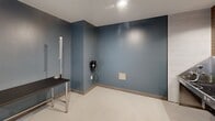 Located at 18750 Delaware St in Huntington Beach, California, this professional pet grooming suite is thoughtfully designed to accommodate all aspects of pet care in a compact yet highly functional space. The interior features a calming palette of muted teal-blue walls complemented by warm earth-toned brick tile accents, creating a welcoming environment for both pets and their owners.

The grooming area is anchored by a dedicated pet bathing station, complete with an elevated tub, integrated steps for easy pet access, and floor drains for efficient water management. Rich terracotta-toned subway tiles line the bathing alcove walls, adding both visual warmth and durable, easy-to-clean surfaces. Stainless steel grooming tables provide sturdy, hygienic workstations throughout the space.

A separate finishing area features blue-painted walls, a wall-mounted professional grooming dryer, and ample maneuvering space. Built-in cabinetry along one wall offers organized storage for grooming supplies and products. Natural light filters in through a window, brightening the space alongside recessed and track lighting overhead.

The light-toned, easy-clean flooring and stainless steel accents throughout reinforce the space's professional, sanitary character, making this suite a well-equipped facility for full-service pet grooming.