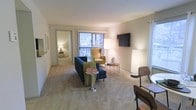 This single-floor residence offers a welcoming and functional living environment highlighted by a well-proportioned living room at its center. The space is finished with wall-to-wall carpeting in a neutral tone that extends throughout, creating a cohesive and comfortable feel underfoot. Soft, light-toned walls contribute to a clean and airy atmosphere throughout the interior.

The living area is the focal point of the space, featuring a wall-mounted flat-screen television and a large sliding glass door fitted with vertical blinds, which allows natural light to filter in while providing access to an outdoor balcony. The room comfortably accommodates a full seating arrangement, including a sofa and accent chair, complemented by ambient floor lamps that enhance the warm atmosphere.

A hallway visible from the living room leads to additional areas of the residence, suggesting a thoughtful and connected layout. The overall design is straightforward and practical, with a neutral color palette that allows for versatile furnishing and personalization. The combination of natural light, outdoor access, and a cohesive interior finish makes this a comfortable and inviting single-floor living space.