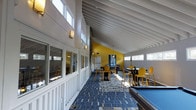 Located in Tate's Creek Village, this single-floor community clubhouse is designed to serve as a vibrant gathering space for residents. The main recreation room is the centerpiece of the facility, boasting soaring vaulted ceilings with exposed white-painted rafters that lend an airy, open feel to the interior. An abundance of clerestory windows lines the upper walls, flooding the space with generous natural light throughout the day.

The room is thoughtfully appointed with multiple functional zones. A billiards area features a full-sized pool table, providing a focal point for recreational activity. Nearby, several high-top dining tables paired with sleek yellow bar-height chairs offer casual seating for socializing or informal meals. A bold yellow accent wall anchors the kitchen end of the room, where a full-sized refrigerator and additional appliances are visible, making the space well-suited for community events and gatherings.

The flooring throughout features a striking blue geometric patterned carpet that adds color and visual interest to the space. Wall-mounted sconces provide warm supplemental lighting alongside the natural light. A stone or brick chimney column rises as an architectural accent, adding a touch of rustic character to the otherwise modern interior. Large windows along the side walls further connect the interior to the surrounding outdoor environment.