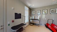 This dedicated Pet Spa is a thoughtfully designed facility built to accommodate all aspects of pet care and grooming. The space is finished with sleek, floor-to-ceiling glossy subway tiles in a soft gray tone that line the grooming area, lending a clean and polished aesthetic. Underfoot, classic white mosaic tile flooring provides a durable, easy-to-clean surface throughout. Natural light pours in generously through multiple large windows fitted with white horizontal blinds, creating a bright and welcoming environment. A professional-grade stainless steel grooming tub with an integrated hose and step stool serves as the centerpiece of the bathing area, while a dedicated grooming table with an attached restraint arm offers a convenient station for styling and finishing. A dog treadmill stored nearby provides an added amenity for pet exercise. Framed pet portraits adorn the tiled walls, adding a charming decorative touch. The space is accessed through a full-glass interior door that connects to a corridor, as well as a direct exterior door leading to the outdoors. Posted pet spa rules ensure a safe and respectful environment for all users. Recessed lighting and well-placed ventilation complete this practical yet stylish pet-friendly amenity.